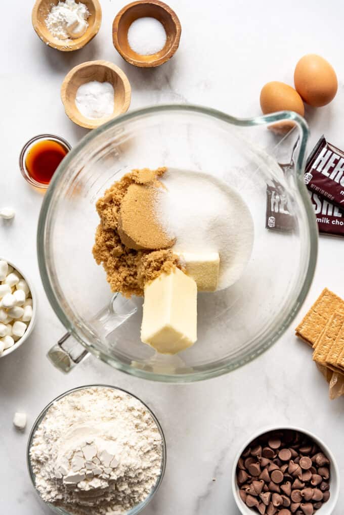 Two sticks of butter, brown sugar, and granulated sugar in a glass mixing bowl surrounded by more ingredients for making cookies.