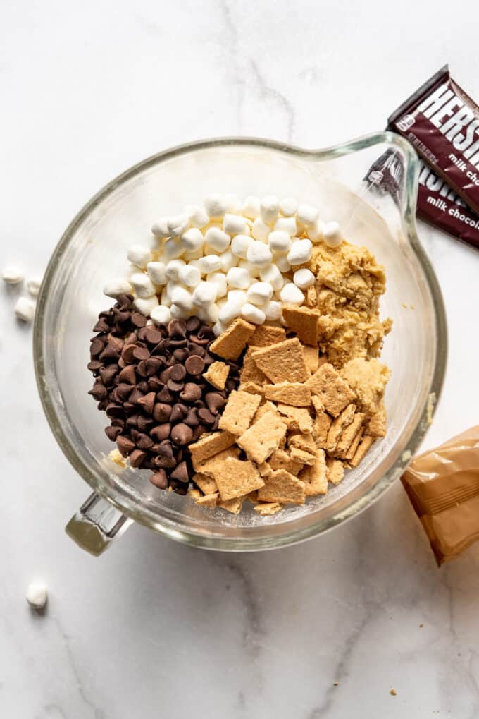 Adding chocolate chips, marshmallows, and graham crackers to a glass bowl.
