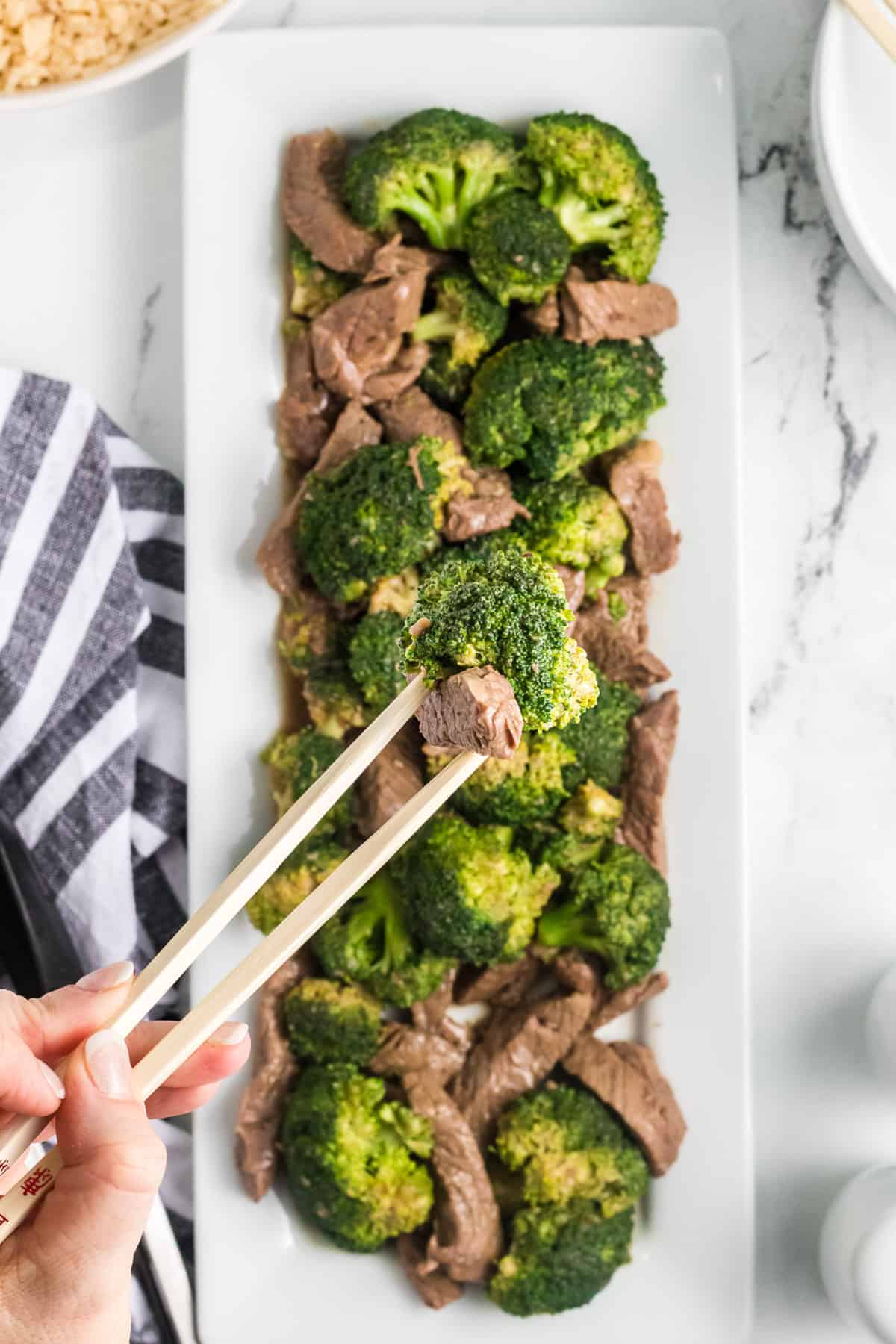 A rectangular serving plate of beef and broccoli with a hand holding chopsticks.