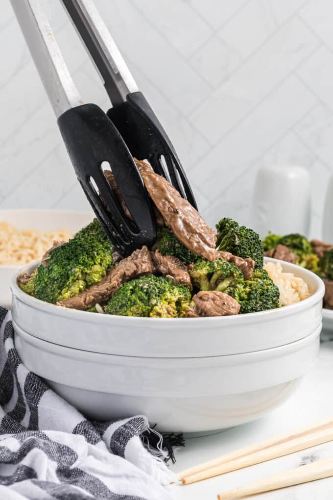 Tongs placing strips of seared beef and broccoli in a bowl over rice.
