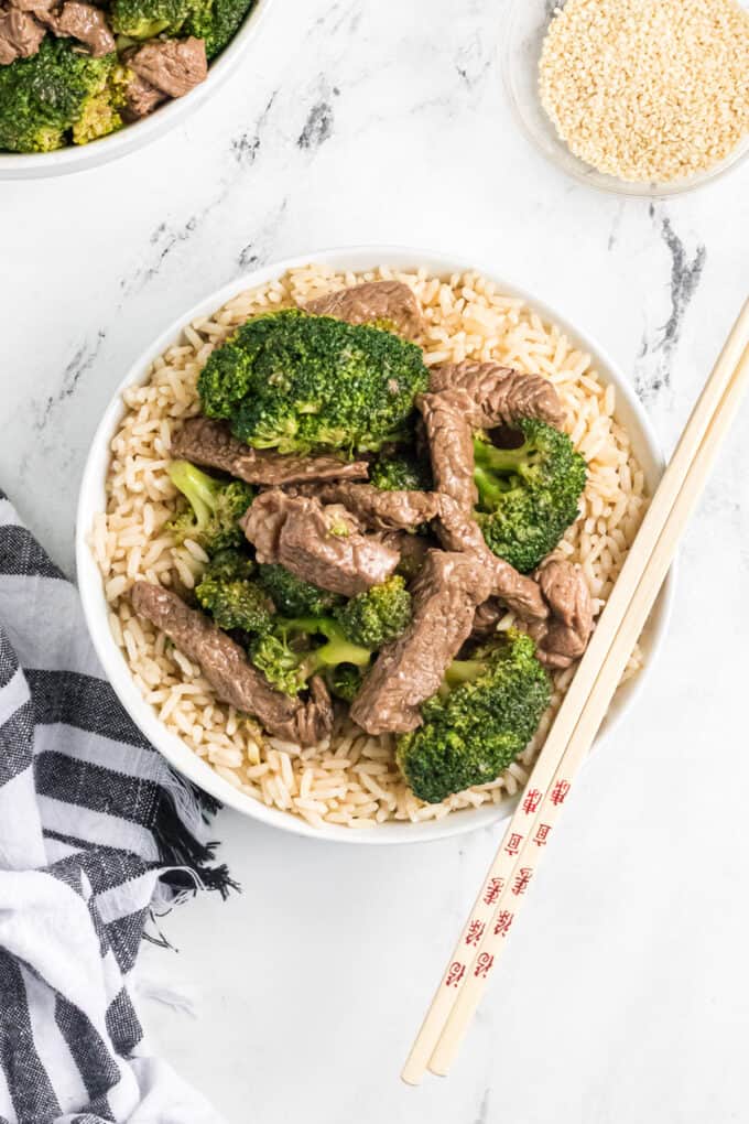 A bowl of beef and broccoli on top of brown rice with chopsticks on the side.