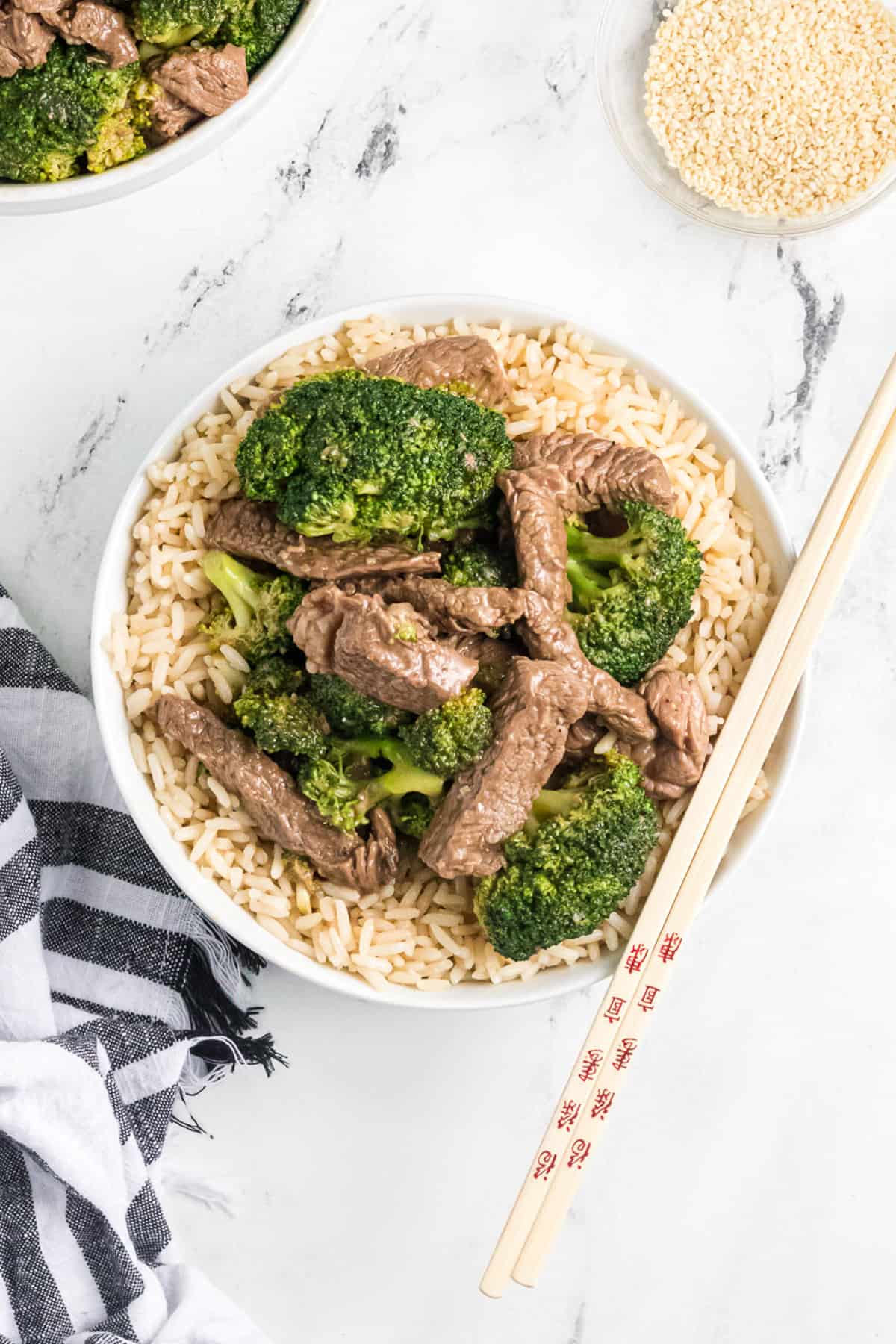 A bowl of beef and broccoli on top of brown rice with chopsticks on the side.