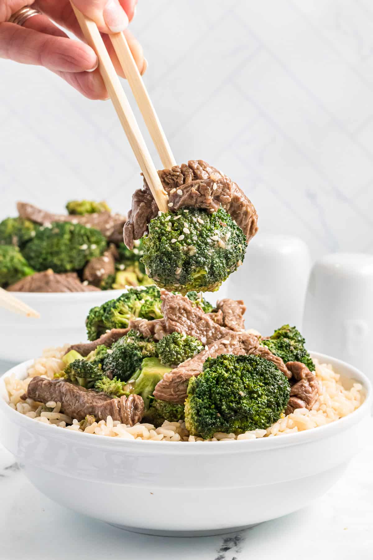 Chopsticks lifting a bite of beef with broccoli from a white bowl.