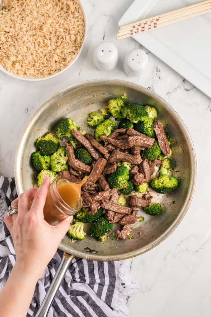 A hand pouring a brown sauce over seared beef and broccoli in a pan.