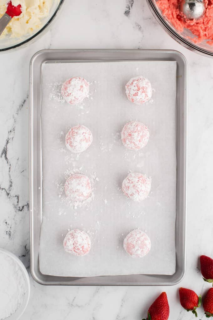 Balls of strawberry cookie dough rolled in powdered sugar on a baking sheet.
