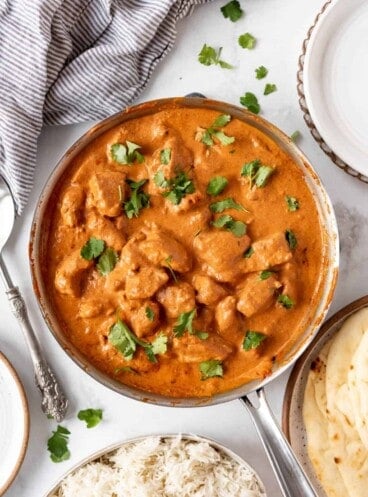 A large skillet of chicken tikka masala next to a bowl of rice and a plate of garlic naan.