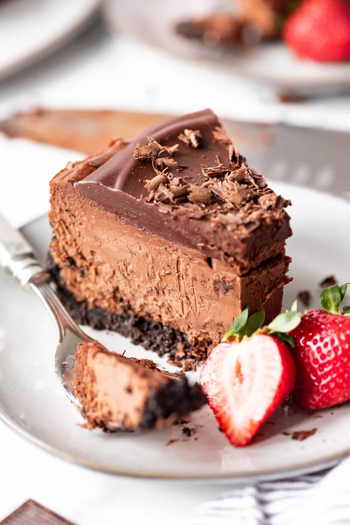 A slice of triple chocolate cheesecake on a plate, with a fork holding a bite-sized portion on the side. 