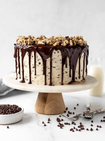 A chocolate chip cookie dough cake with chocolate ganache on a marble cake stand surrounded by mini chocolate chips.