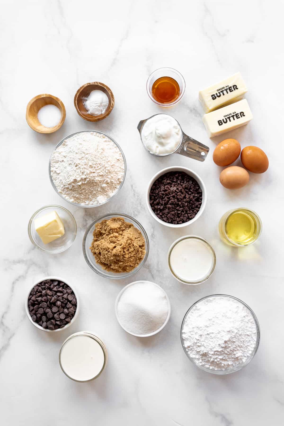 Ingredients in separate bowls on a white marble surface for making chocolate chip cookie dough cake.