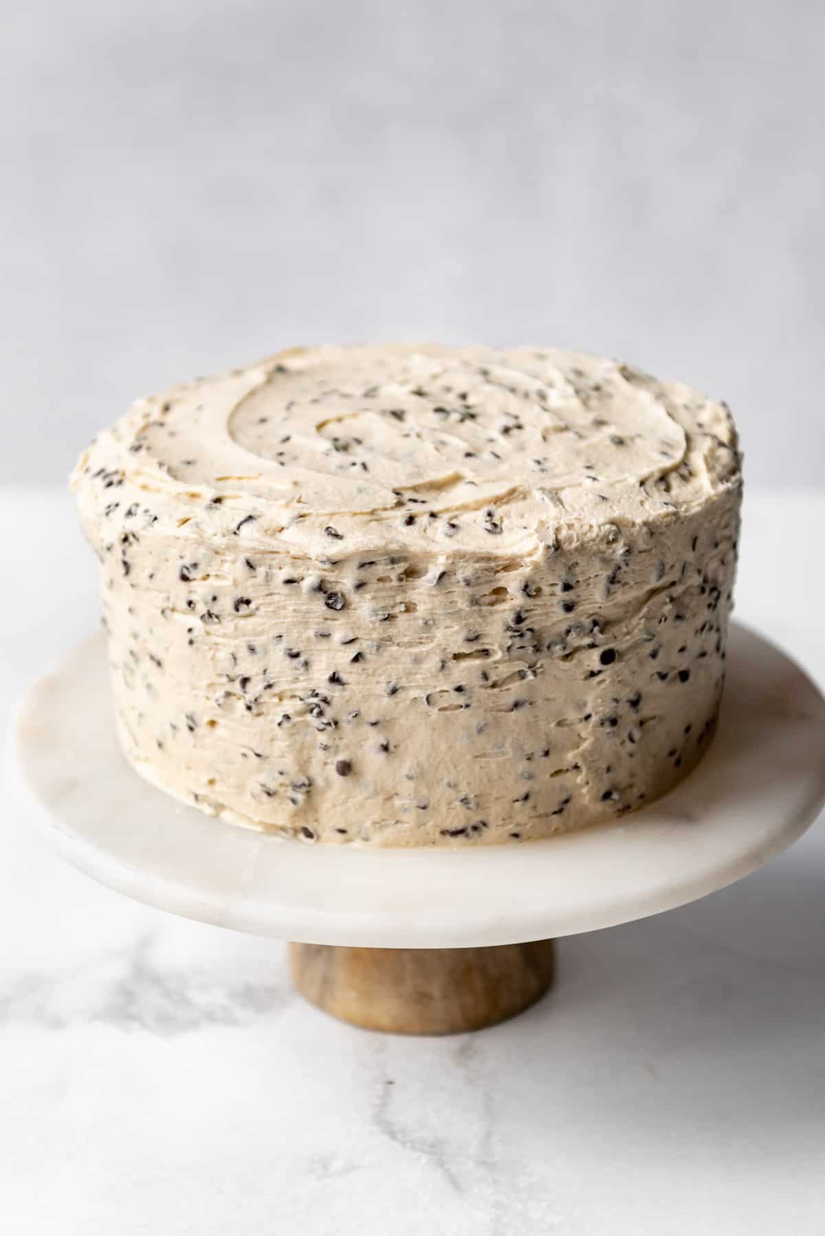 A cake frosted with chocolate chip cookie dough frosting on a white marble cake stand.