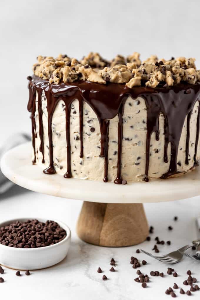 A chocolate chip cookie dough cake with chocolate ganache on a marble cake stand surrounded by mini chocolate chips.