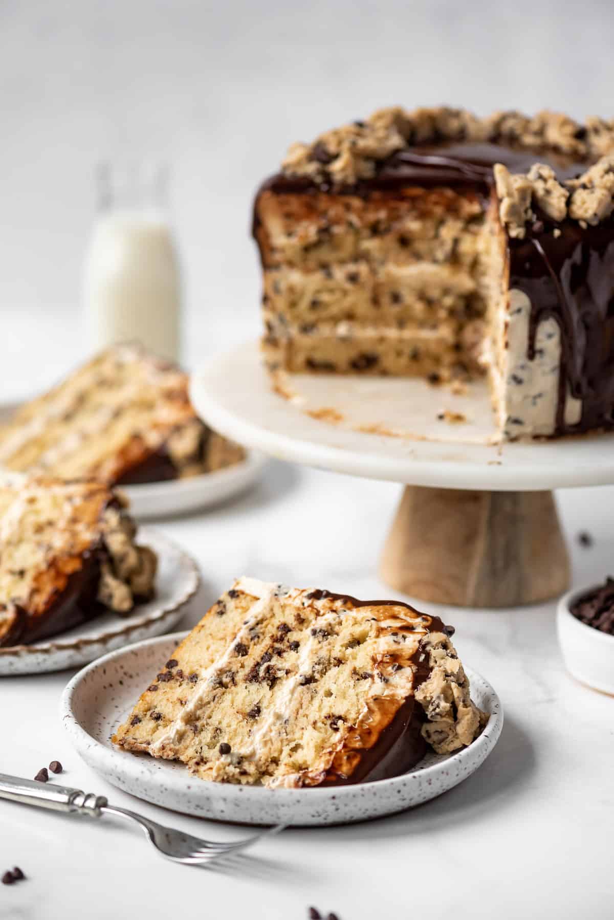 Slices of chocolate chip coookie dough cake on plates next to the rest of the cake on a cake stand.