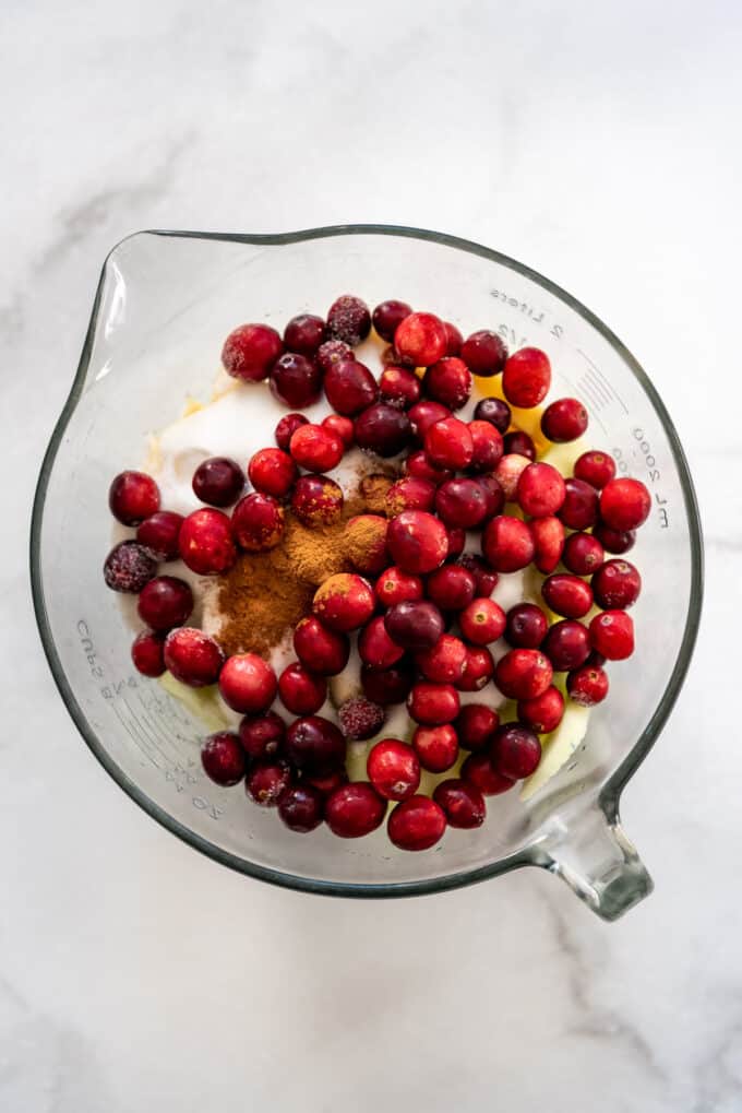Fresh cranberries on top of sliced Granny Smith apples, sugar, flour, and cinnamon in a glass bowl.