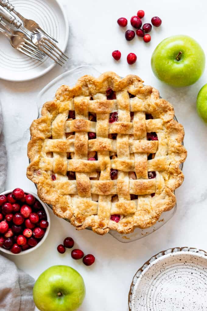 A lattice crust topped pie next to a bowl of fresh cranberries and granny smith apples.