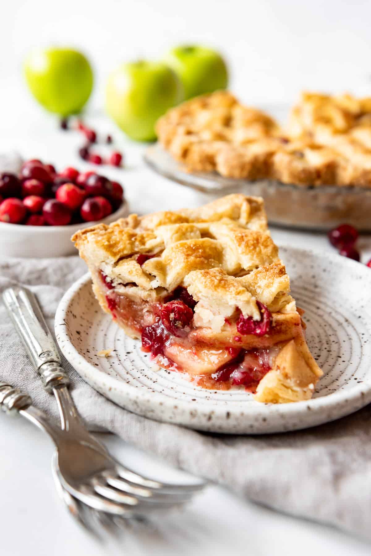 A piece of cranberry apple pie on a plate in front of the rest of the pie and a bowl of fresh cranberries.