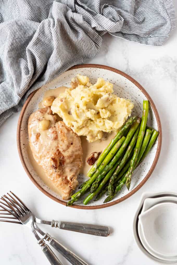 A plate of creamy garlic chicken, mashed potatoes, and asparagus, next to forks and a linen napkin on a white marble surface.