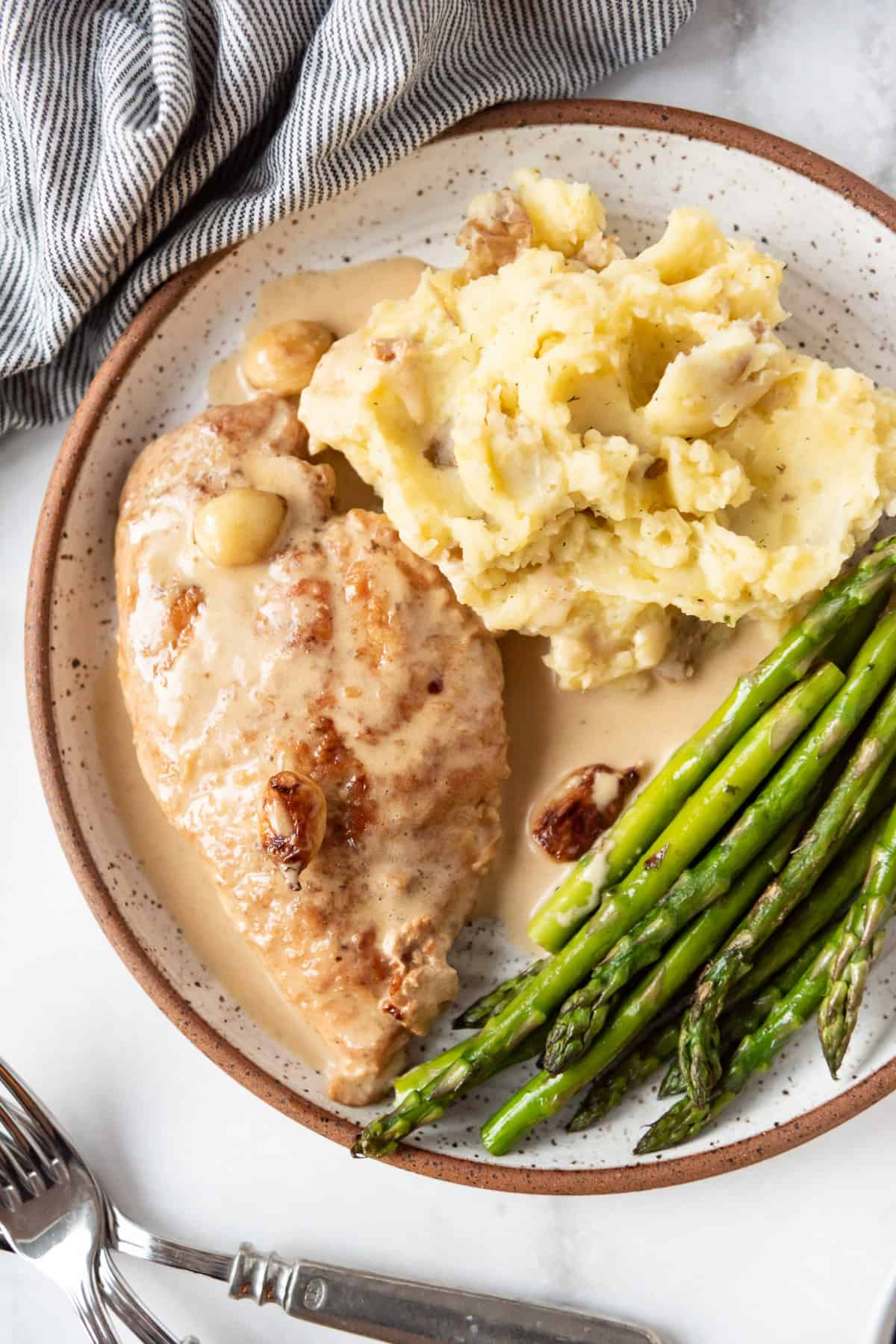 Pan-seared chicken breast cooked in a creamy garlic slice with whole garlic cloves being served with mashed potatoes and asparagus.