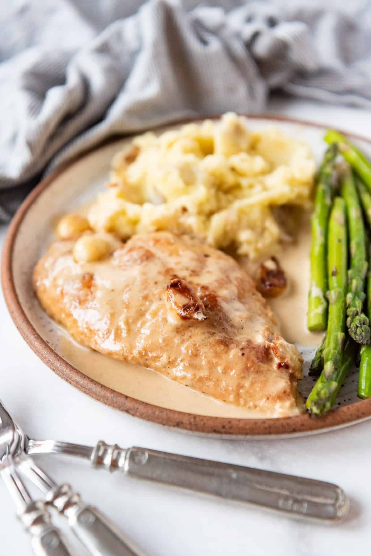 A piece of creamy garlic chicken on a plate with mashed potatoes and asparagus in front of a linen napkin.
