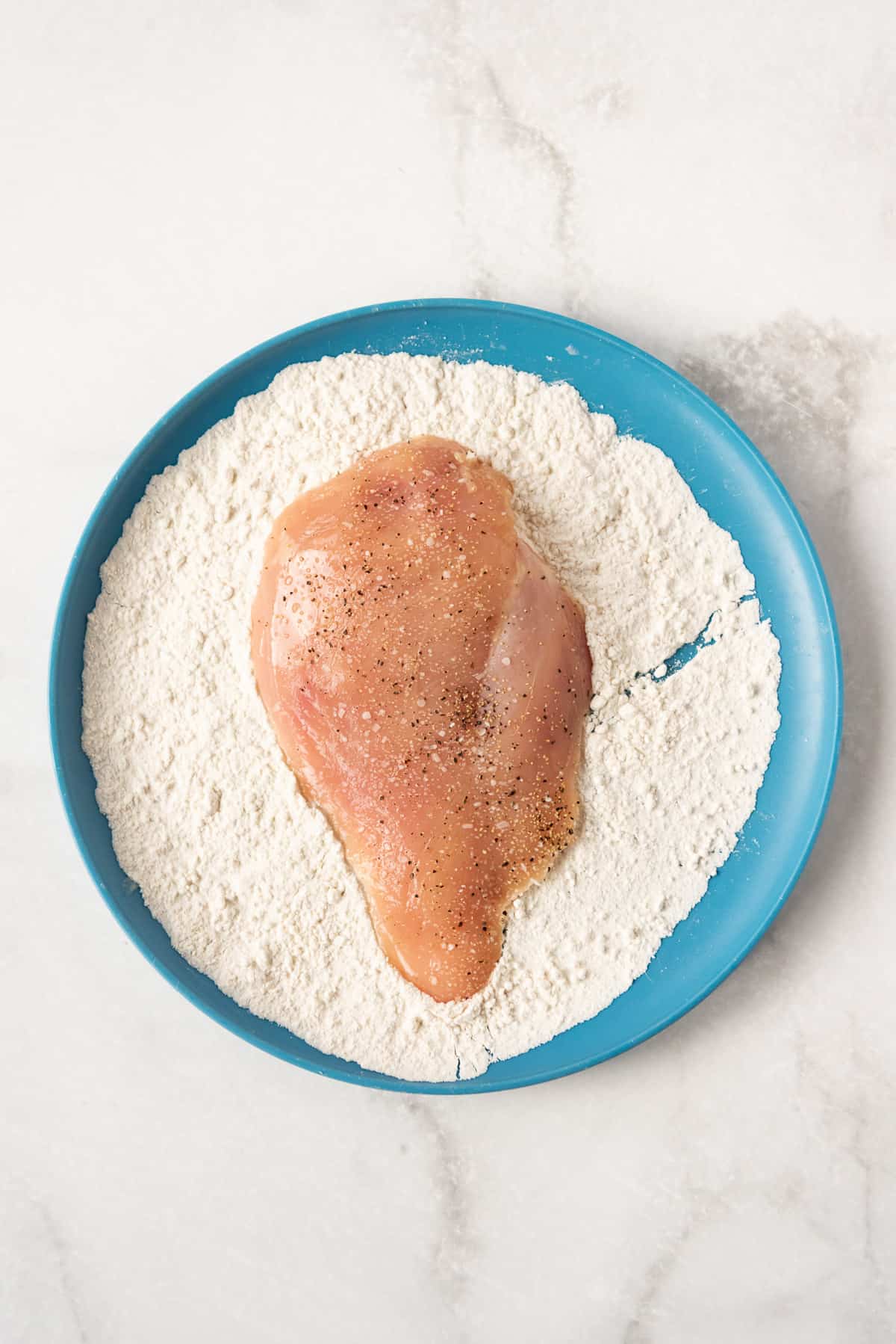 Dredging seasoned chicken cutlets in flour on a blue plate.