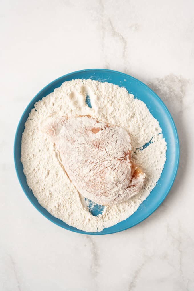 Dredging seasoned chicken cutlets in flour on a blue plate.