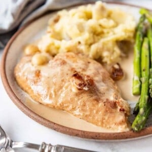 A chicken breast in creamy garlic sauce on a plate with mashed potatoes and asparagus.