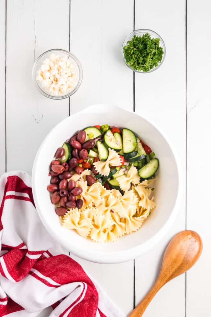 Combining pasta, kalamata olives, and sliced cucumbers in a large bowl.