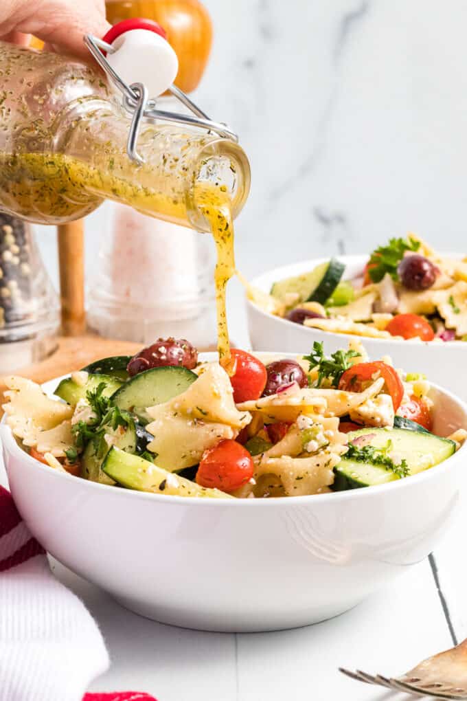 Pouring Greek dressing over a bowl of pasta salad.