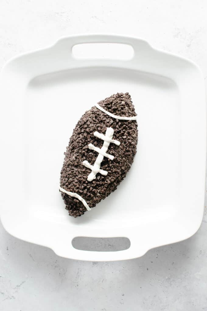 A mini chocolate chip-covered dessert cheese ball decorated to look like a football on a white platter.