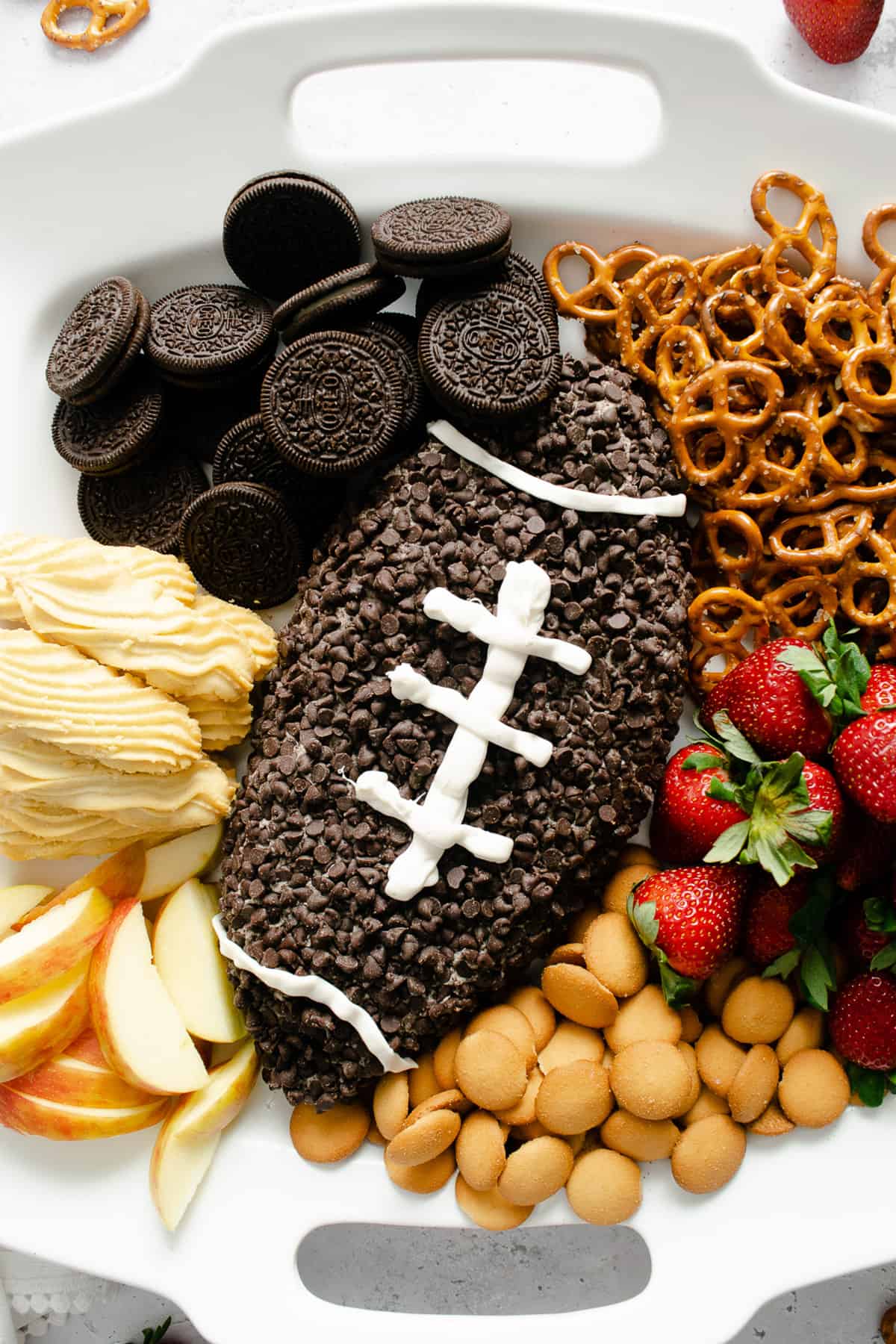An Oreo cheesecake cheese ball decorated like a football for Game Day on a platter with pretzels, strawberries, and cookies for dipping.