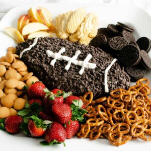 An Oreo cheesecake cheese ball decorated like a football for Game Day on a platter with pretzels, strawberries, and cookies for dipping.