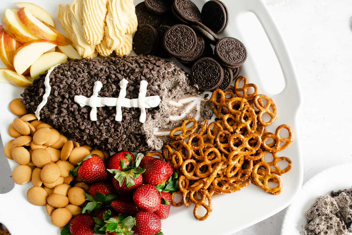 A football shaped dessert cheese ball with a few servings removed on a serving platter with Oreos, pretzels, strawberries, and other cookies.