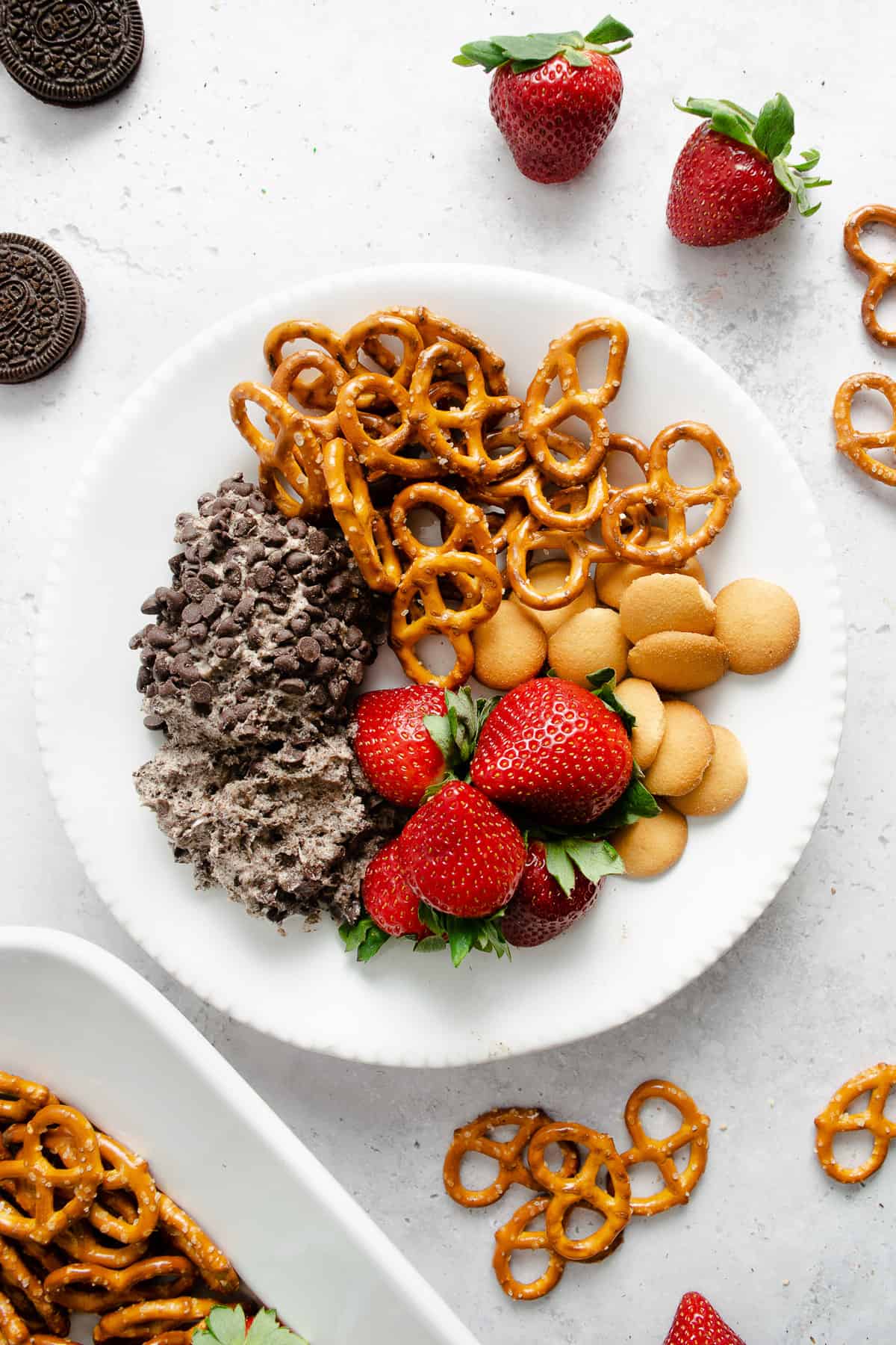 A white plate with a serving of cookies & cream cheesecake dip on it with pretzels, mini Nilla wafers, and strawberries.