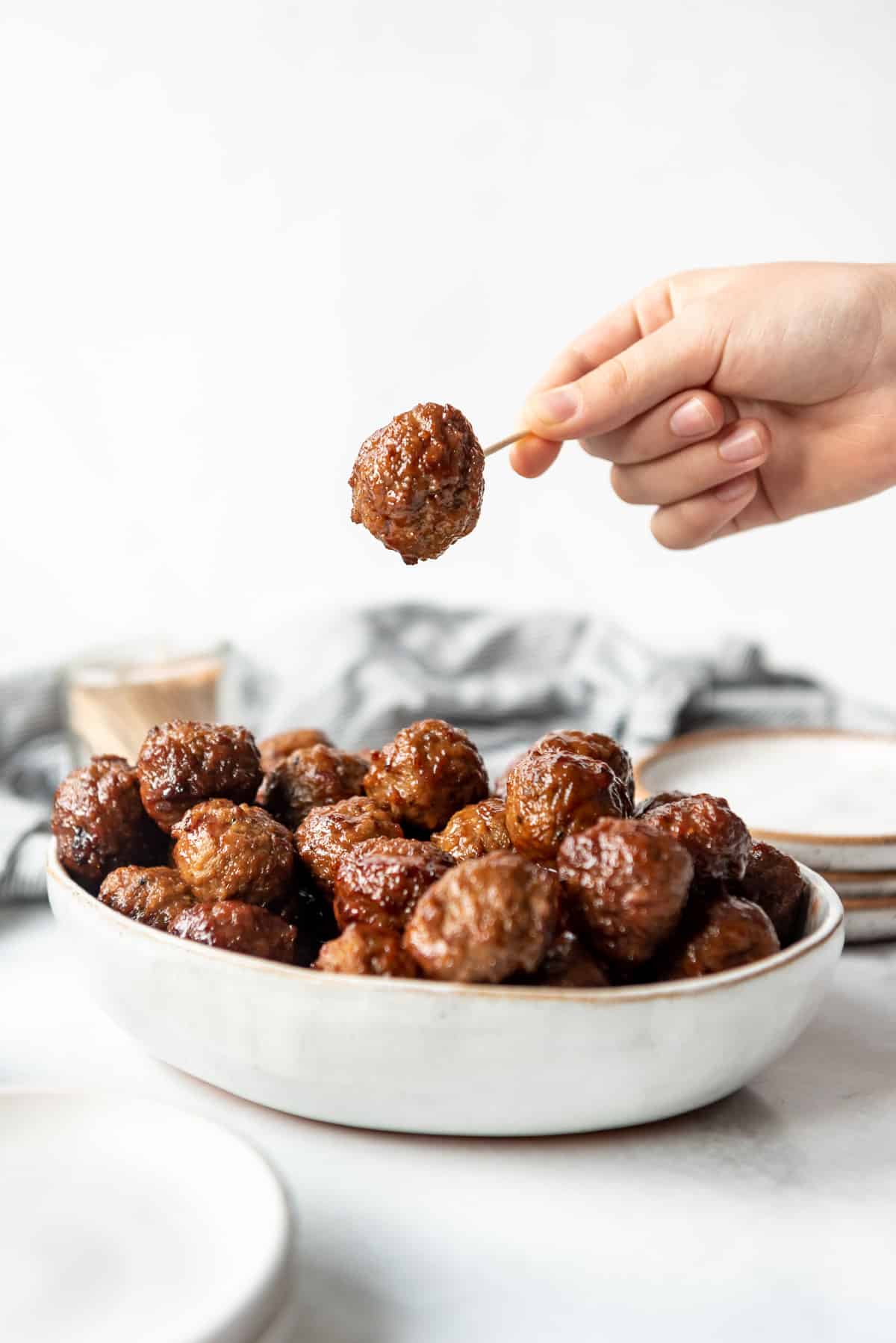 A hand lifting a grape jelly meatball on a toothpick over a bowl of the meatballs.