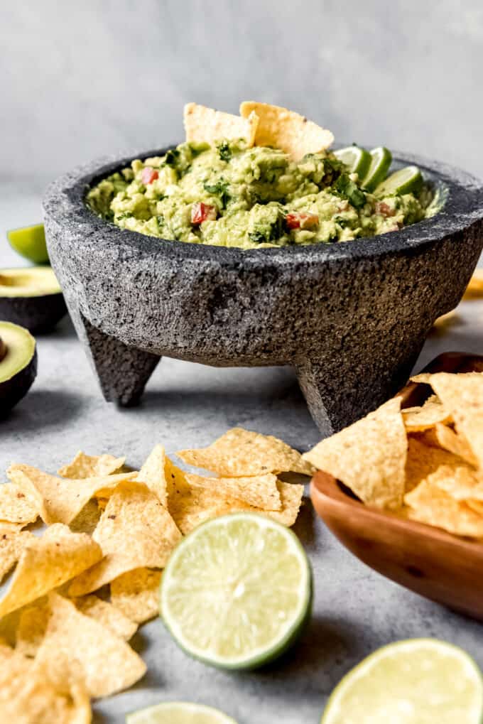 An image of a molcajete stone mortar and pestle filled with homemade guacamole with chips and limes on the side.