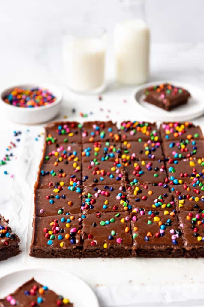 Rows of brownies with chocolate frosting and rainbow coated chocolate chips sprinkled on top in front of a bowl of sprinkles and a couple of glasses of milk..