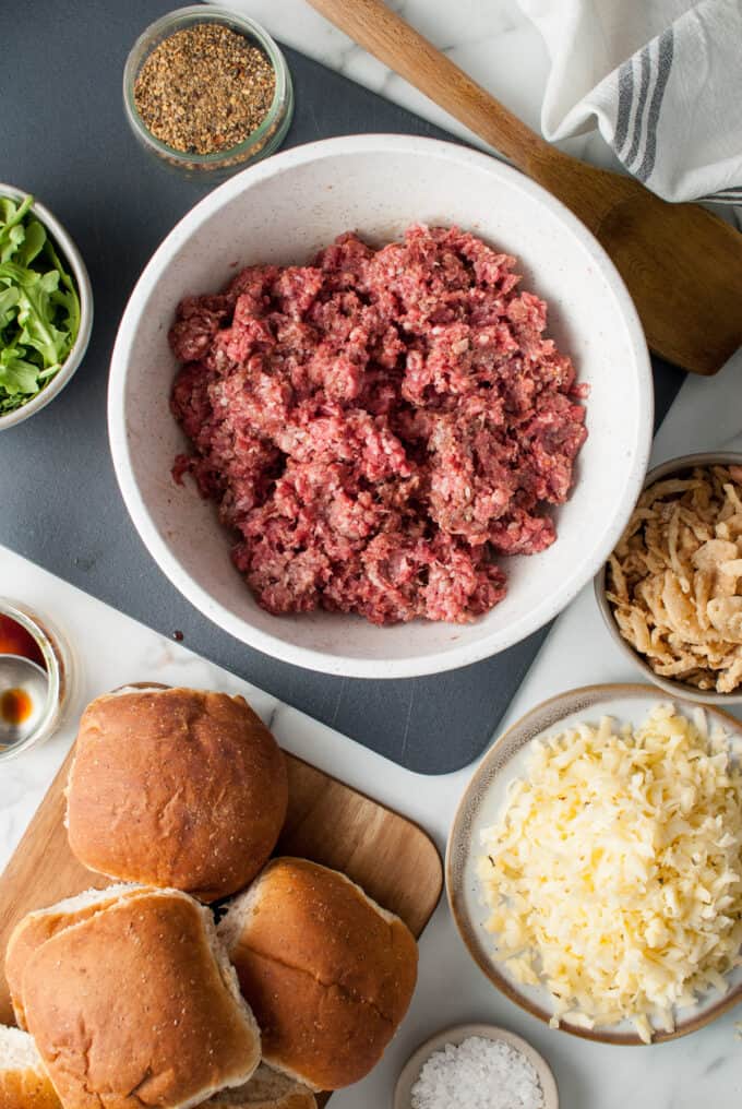 Seasoned ground beef in a mixing bowl surrounded by burger buns, shredded cheese, and other ingredients.