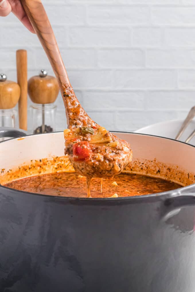 A wooden serving ladle lifting up a scoop of lasagna soup over a grey pot.