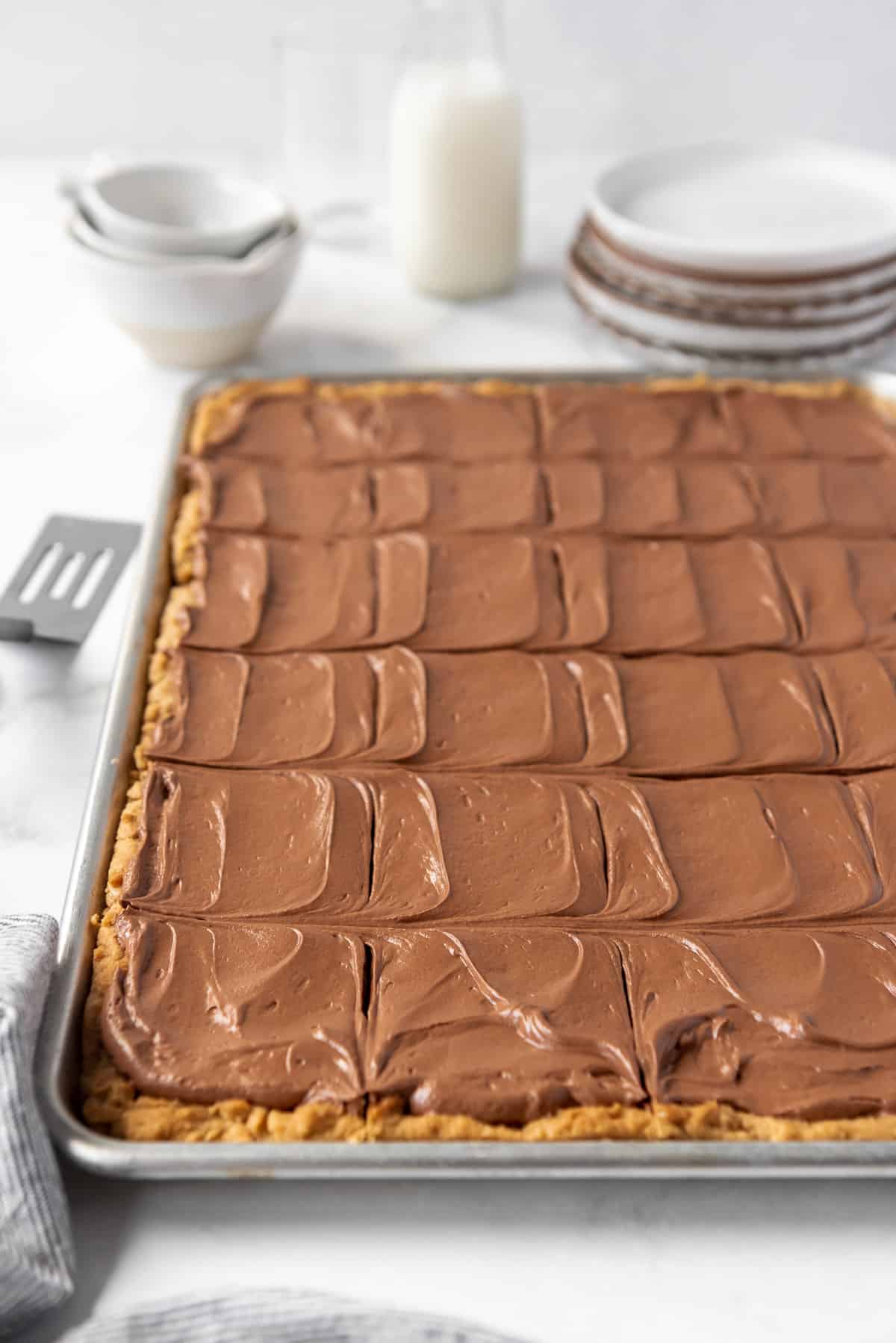 A try full of peanut butter oatmeal bars with chocolate frosting in front of a stack of plates and a glass of milk.