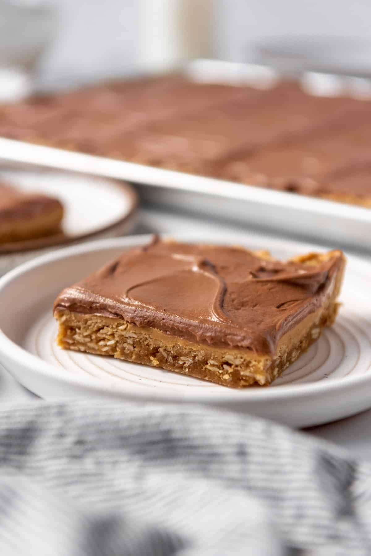A lunch lady peanut butter bar with chocolate frosting on a white plate in front of more bars on a baking sheet behind it.