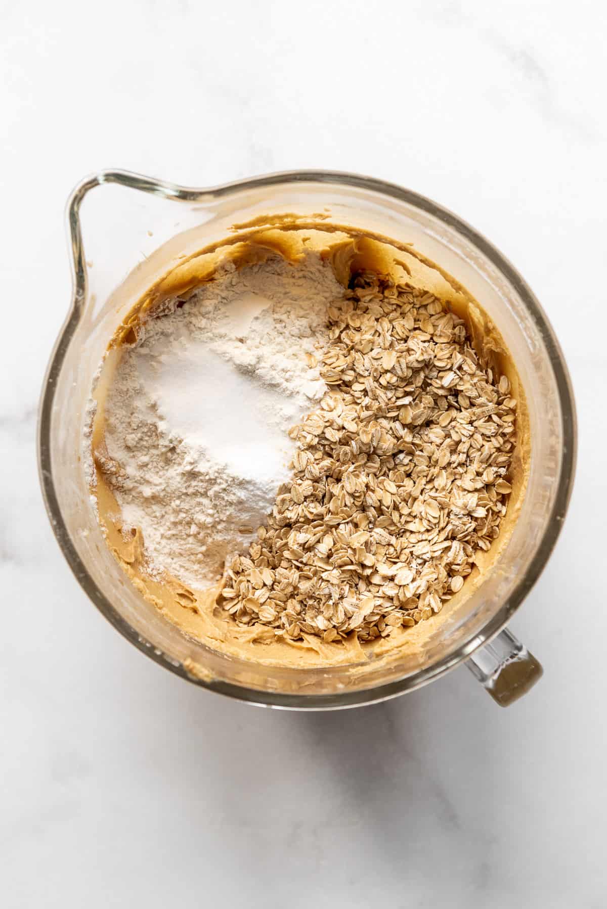 Adding flour, salt, baking soda, and old-fashioned oatmeal to a peanut butter cookie dough base in a glass mixing bowl.