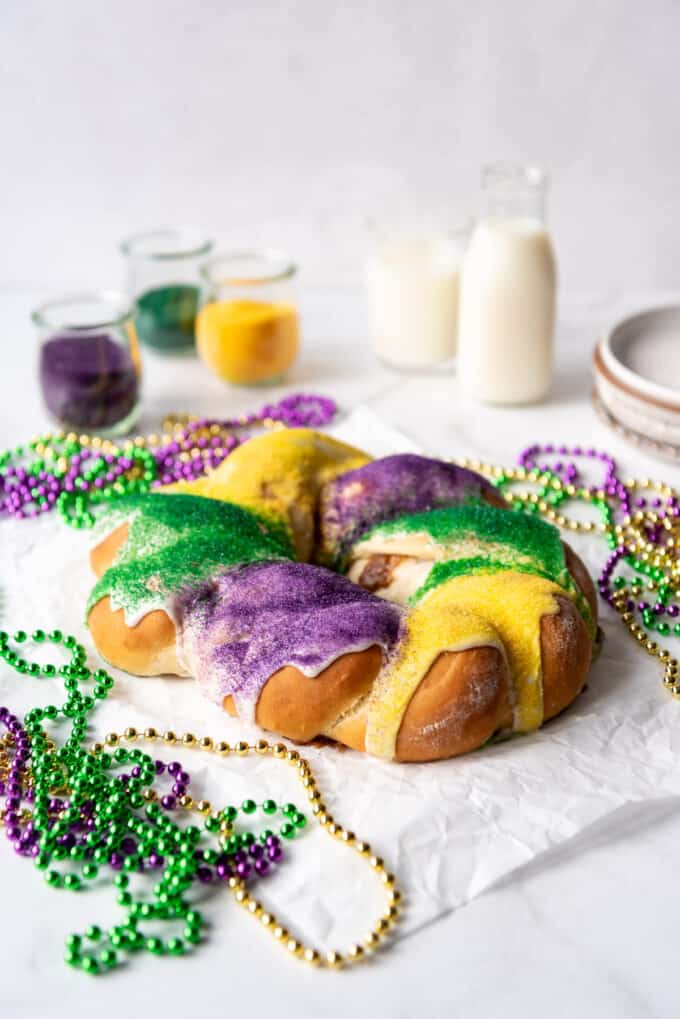 A homemade King Cake surrounded by Mardi Gras beads in front of glasses of milk and jars of colored sugar.
