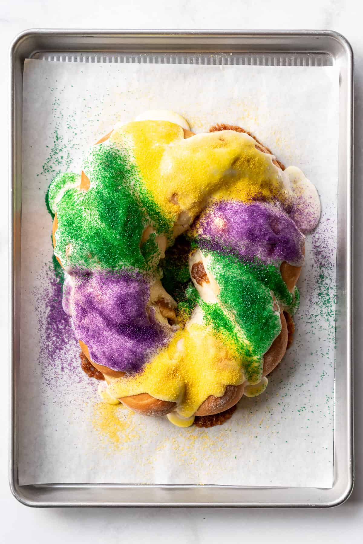 Adding green, purple, and yellow sprinkles to the top of a frosted king cake on a baking sheet.