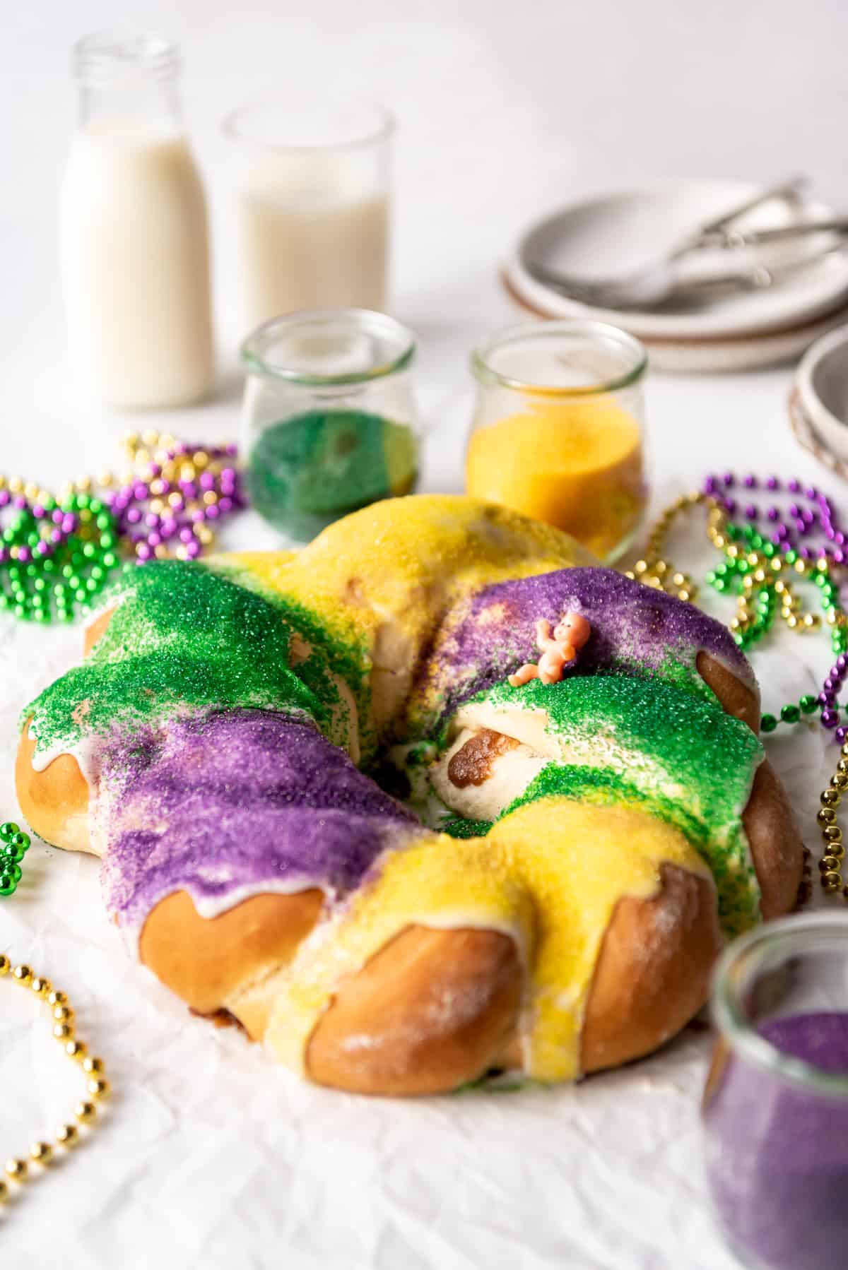 A homemade king cake covered with colored sugar in front of glasses of milk and plates.