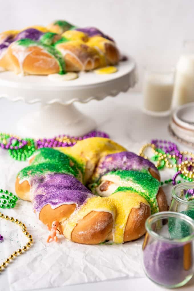 A king cake covered in green, purple, and gold sprinkles in front of another cake on a cake stand.