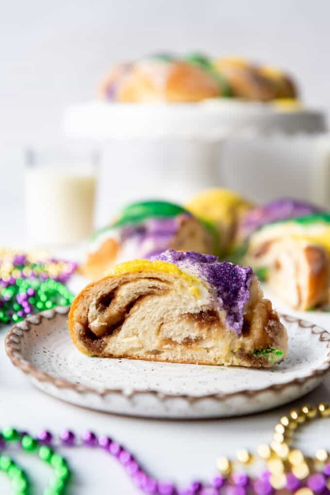 A slice of cinnamon filled Mardi Gras king cake on a plate with a cake stand in the background.