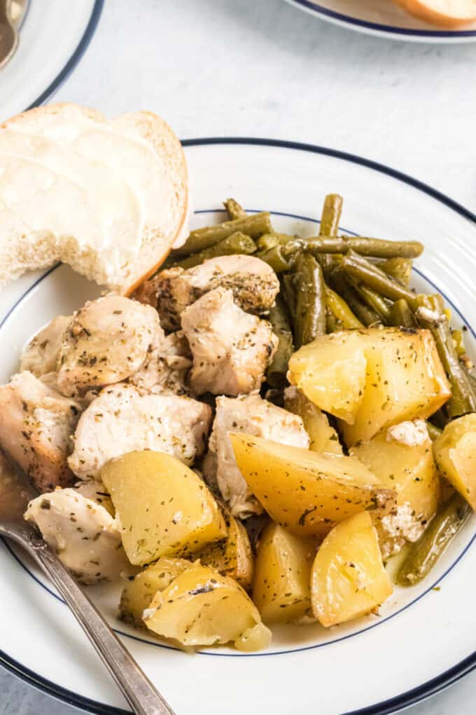 Cooked chicken, potatoes, and green beans on a plate with a piece of buttered bread.