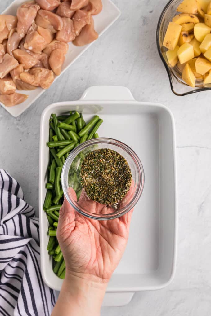 A hand holding a small glass bowl of seasonings over a baking dish with green beans in it.