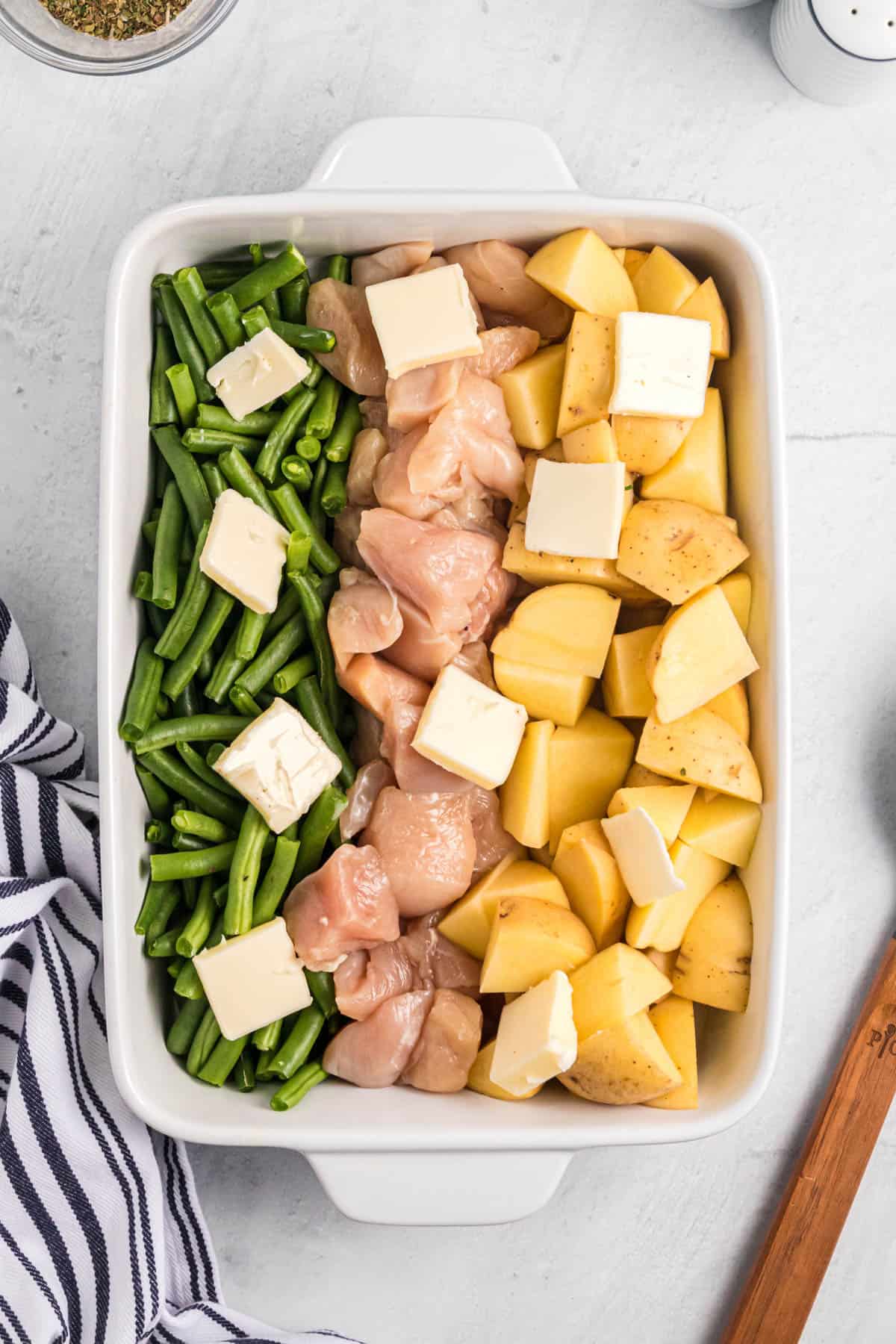 Adding pats of butter on top of raw green beans, chicken chunks, and potatoes in a pan before roasting.