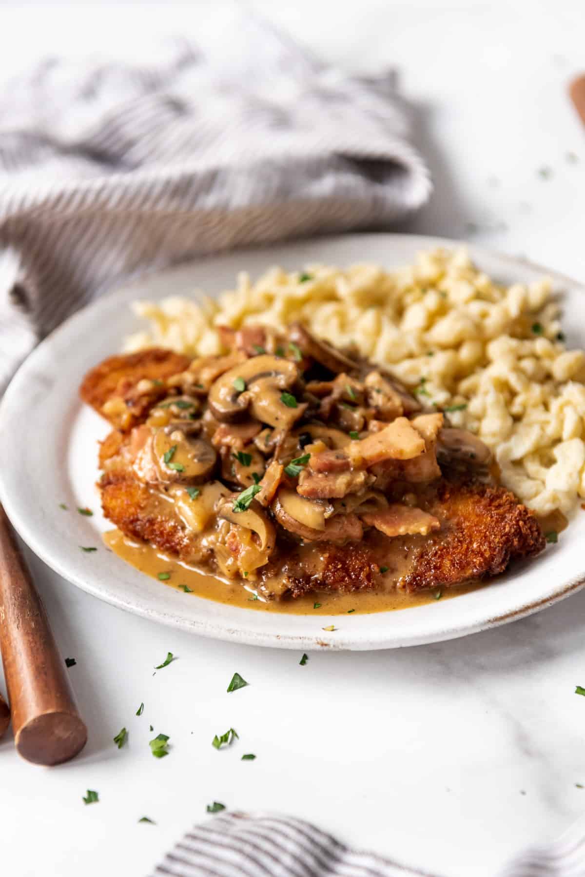 A serving of pork schnitzel topped with brown mushroom gravy and bacon on a plate with noodles.