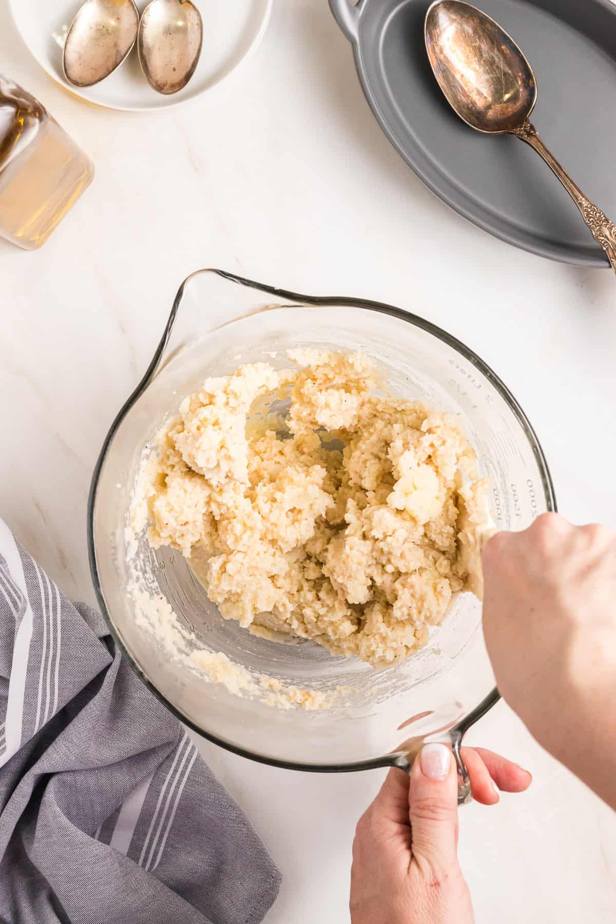 Hands stirring a garlic almond paste into mashed potatoes in a glass bowl.